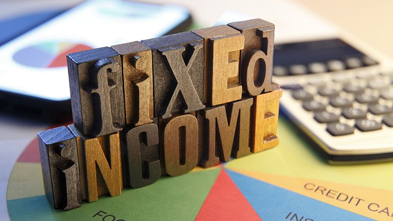 Letterpress blocks spell out "Fixed Income" as they rest on top of a pie chart and next to a calculator and a mobile device.