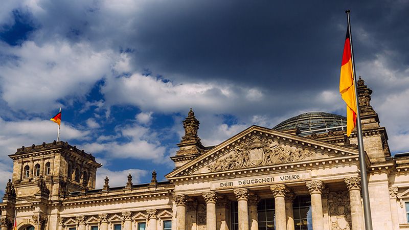 Striking image of the Reichstag building in Berlin under a dramatic cloudy sky. The German flag flutters proudly, encapsulating a sense of national pride and history.